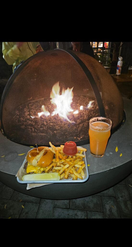 A metal tray with a cheeseburger, french fries, ketchup, and a pickle spear sits on the edge of a large, circular fire pit. To the right of the tray is a glass of hazy, light-colored beer. Inside the fire pit, which is covered by a mesh dome, bright orange flames are visible. The scene is outdoors at night, lit by the fire.