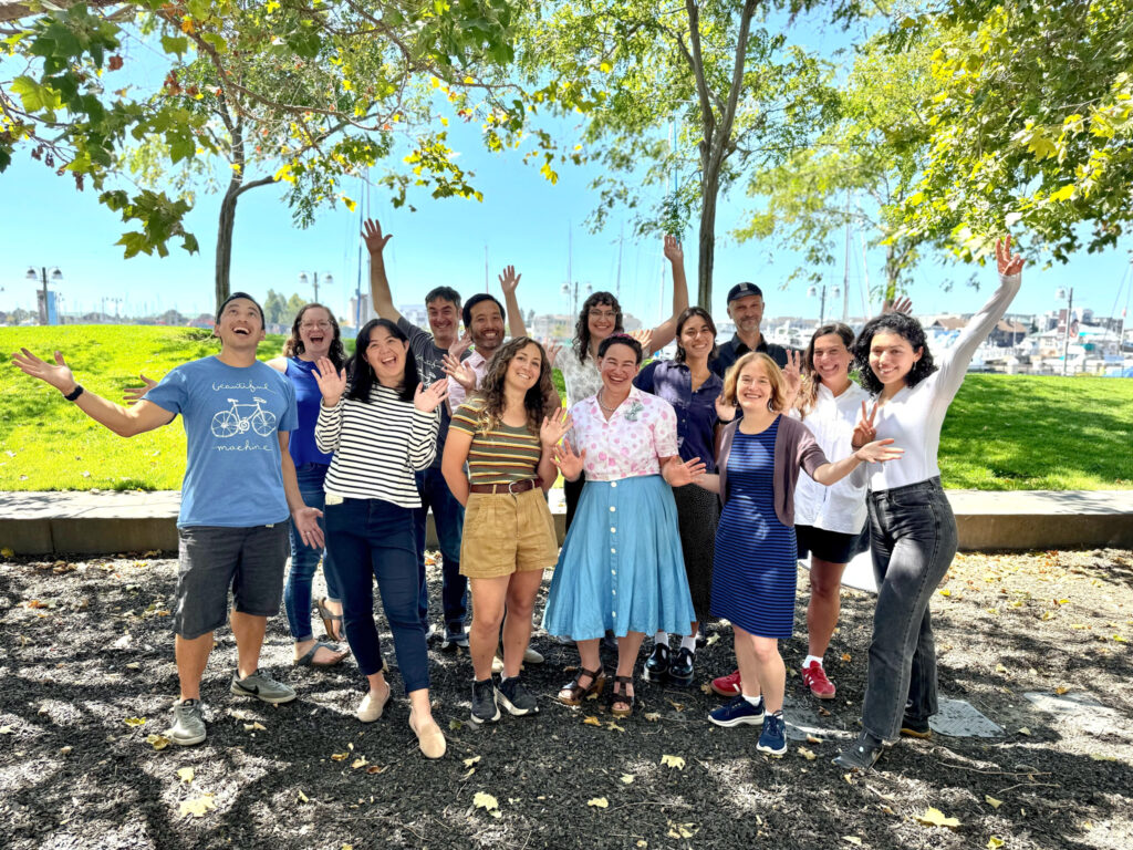 Bike East Bay's 13 person staff smiles and throws their hands in the air in celebration, with trees and a marina in the background.