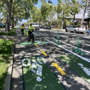 photo of two people biking in a tree-shaded, 2-way post-separated cycletrack as a temporary pop-up installation on Ninth St in Berkeley
