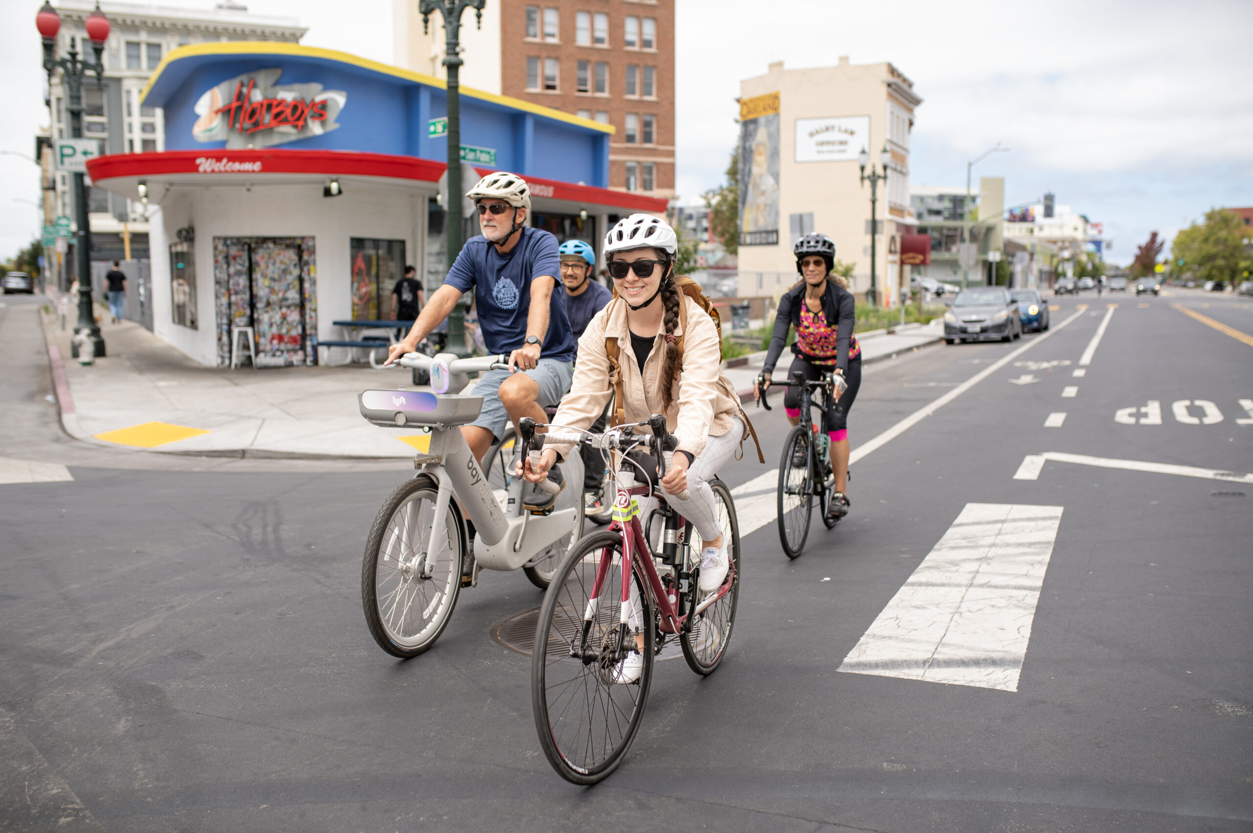 A man, a woman, and a young woman are riding bicycles in a city. The man is on a light gray bike-share bicycle with an electric motor. The young woman in front of him is on a red road bike. Another woman on a black bike is riding behind them. They are all wearing helmets. They are biking down a street with a building and a 'Welcome' sign in the background. The street has a yellow bike lane and white painted lines. The sky is partly cloudy.