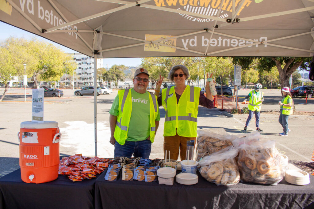 Two event volunteers, a man and a woman, both wearing bright neon yellow safety vests, are standing behind a vendor table covered in snacks under a tent. The man on the left is wearing a San Francisco Giants cap and a green shirt, smiling at the camera. The woman on the right is also smiling and holds up two small bottles of liquid. The table displays large bags of bagels, individual packages of cookies, small cups of dips, and red bags of snacks next to a large orange Igloo water cooler. The event takes place outdoors in a paved lot on a sunny day with trees and buildings in the background.