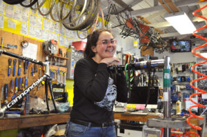 Hannah is smiling as she leans on a bike rack, standing in a bike mechanic shop with tools and bike parts all around.