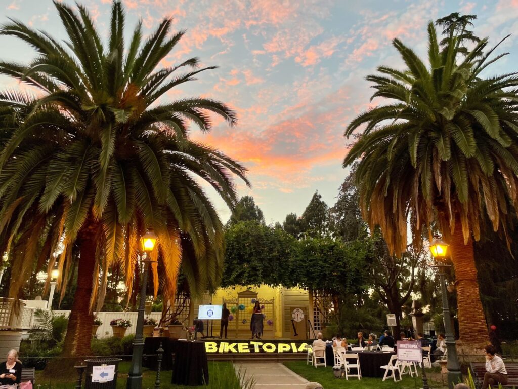 An evening shot of an outdoor event with a stage and seating. The sky is a gradient of orange, pink, and blue at sunset. Two large palm trees with fronds framing the top of the photo are lit by warm lamps. People are sitting at tables in the grassy area to the right, and two people are on a stage with a screen and a sign that says **BIKETOPIA**. There are a few trees in the background, and the overall atmosphere is peaceful.