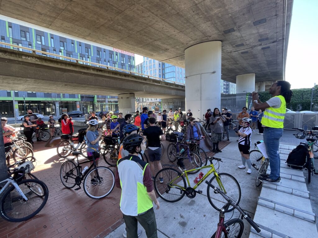 A large group of cyclists listen to instructions from Ride Leader Phil; all are standing with bikes at a BART station