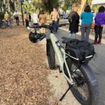 A photo of a parked e-bike along the side of a smoothly paved multi-use trail, with ribbon cutting participants gathering in the background