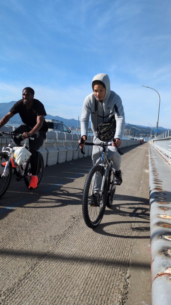 Two people are cycling side-by-side across a bridge on a sunny day